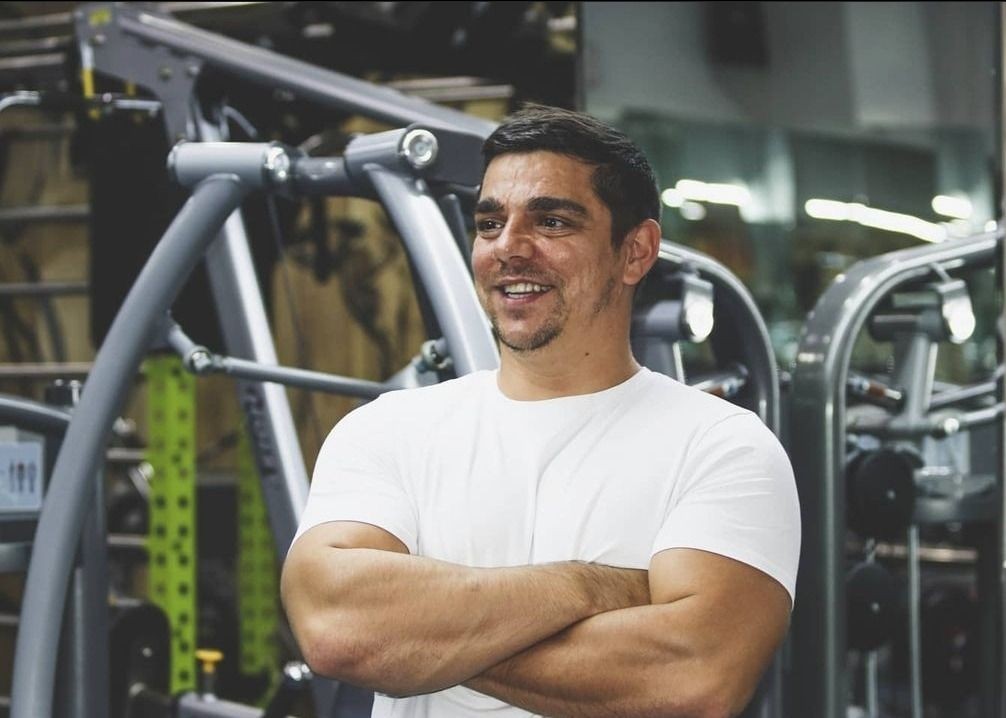 Man in white t-shirt with folded arms standing in a modern gym equipped with various exercise machines.
